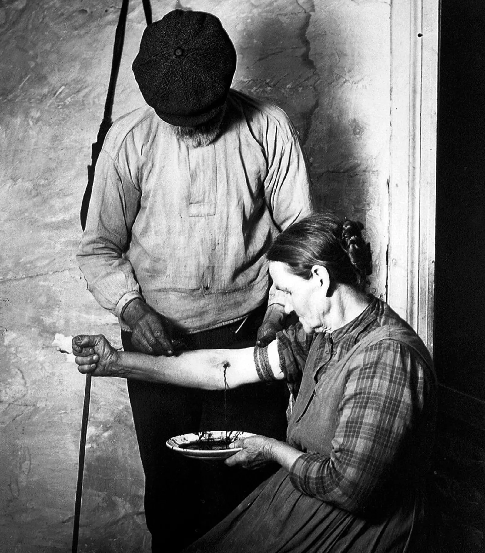 A photograph of a standing man in a cap holding out his bare forearm, which is bleeding into a shallow dish held by a seated woman.